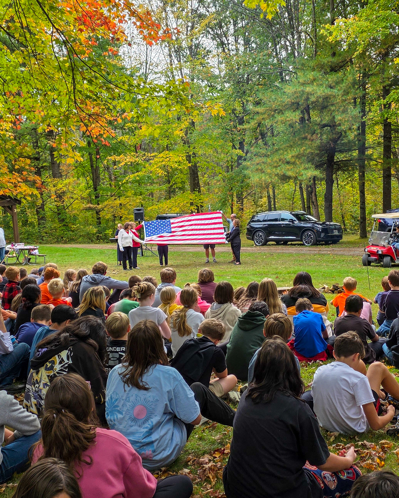 Speakers unfurling a large American flag outdoors in front of a seated audience of students in a park.