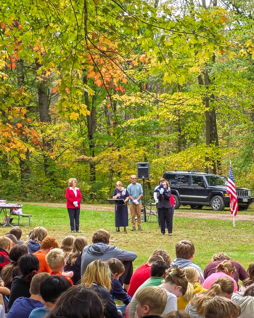 A group of students sitting outdoors in a forest setting, watching speakers holding a folded American flag next to a flag on a pole.