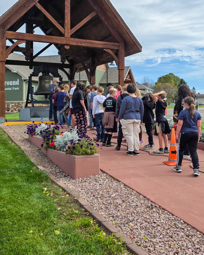 A line of students standing under a wooden gazebo structure holding a rope attached to a large ceremonial bell.