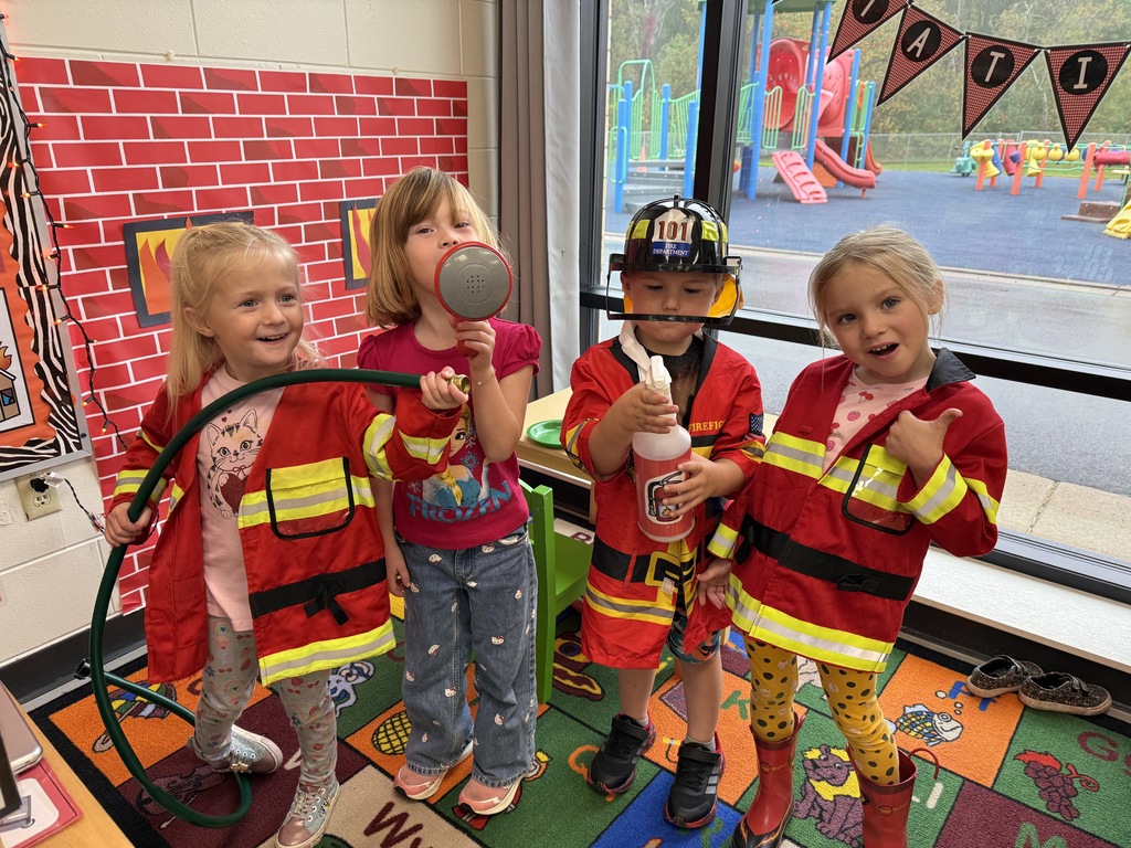 Four 4K students dressed in firefighter coats and helmets pose together in their classroom while holding play hoses, a spray bottle, and a megaphone in front of a pretend brick wall backdrop.