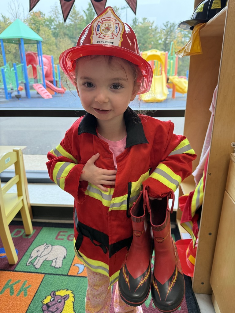 A young student smiling in a firefighter costume holds up a pair of red rubber boots while standing on a colorful classroom alphabet rug.