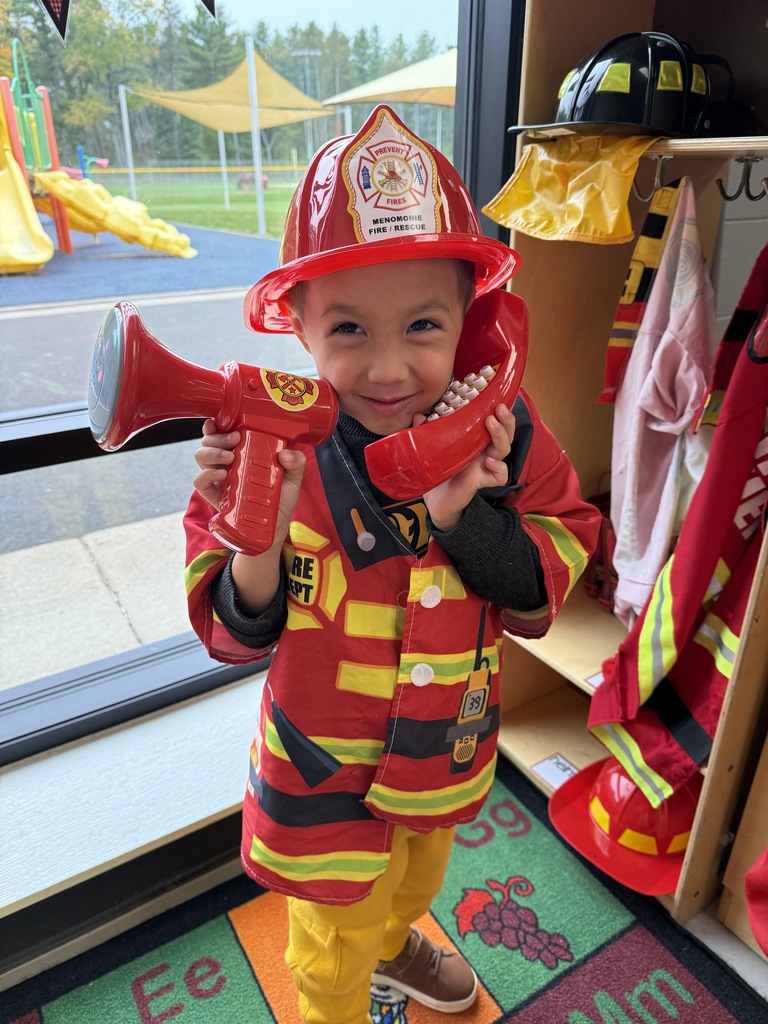 A smiling 4K student dressed in a firefighter jacket and red helmet holds up a red toy megaphone and telephone receiver inside the classroom.