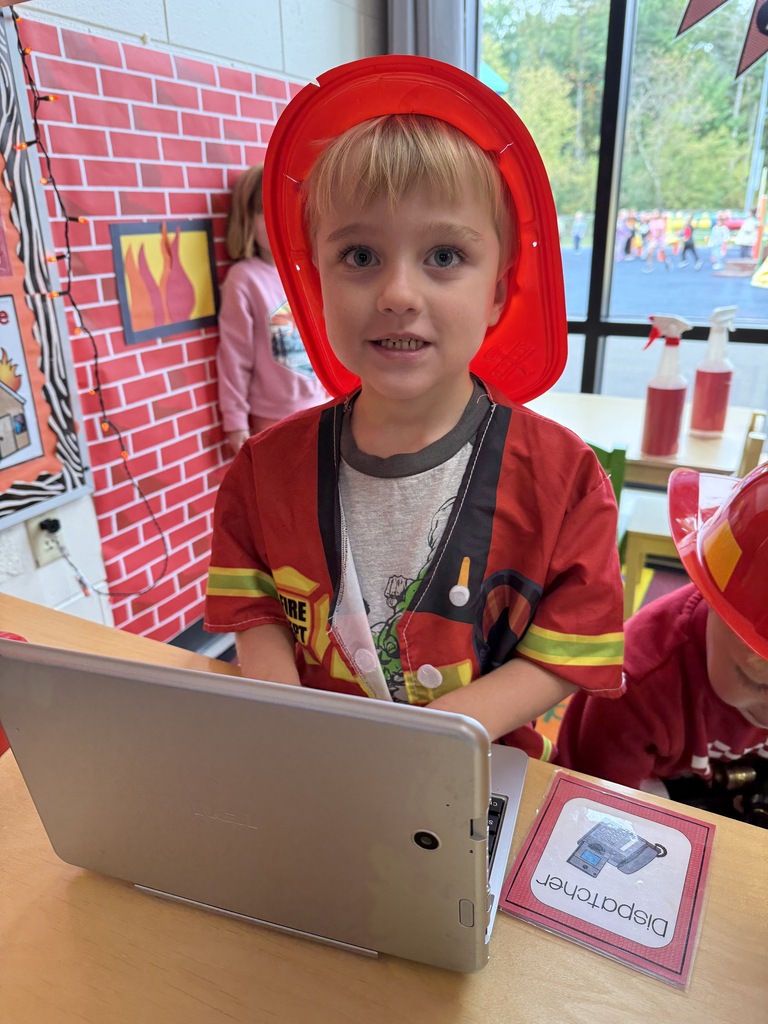 A young 4K student wearing a red firefighter helmet and jacket sits behind a tablet and a "Dispatcher" sign in a pretend classroom fire station setup.