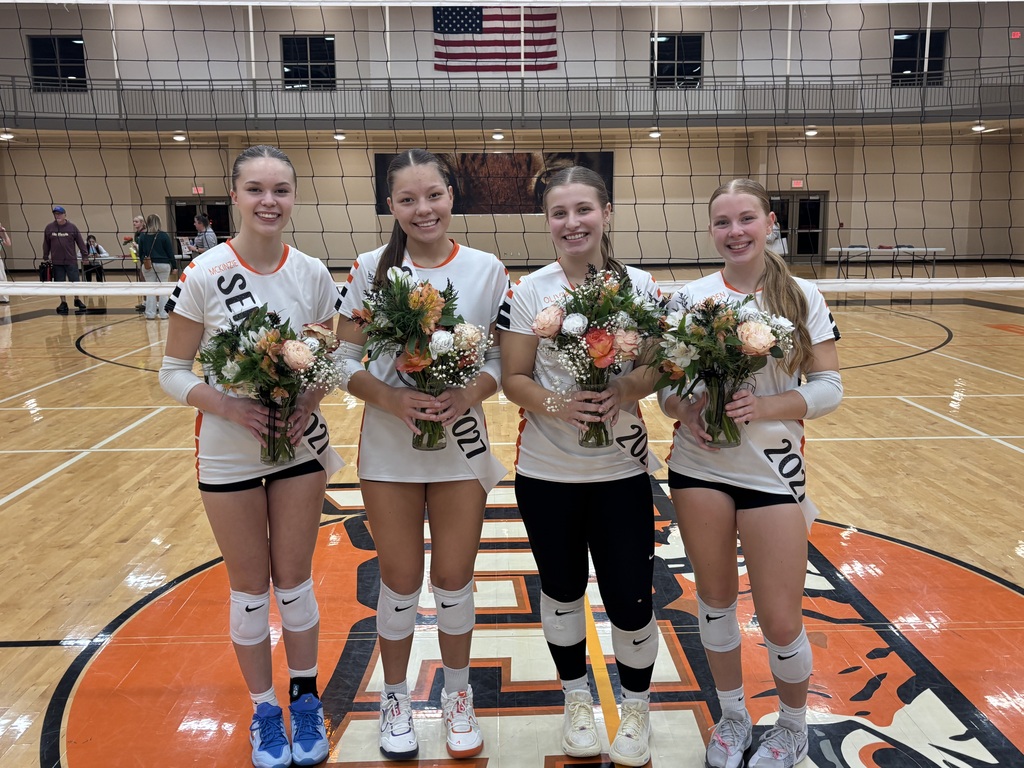Four Black River Falls volleyball seniors wearing senior sashes pose together on their home court holding bouquets of flowers on Senior Night.