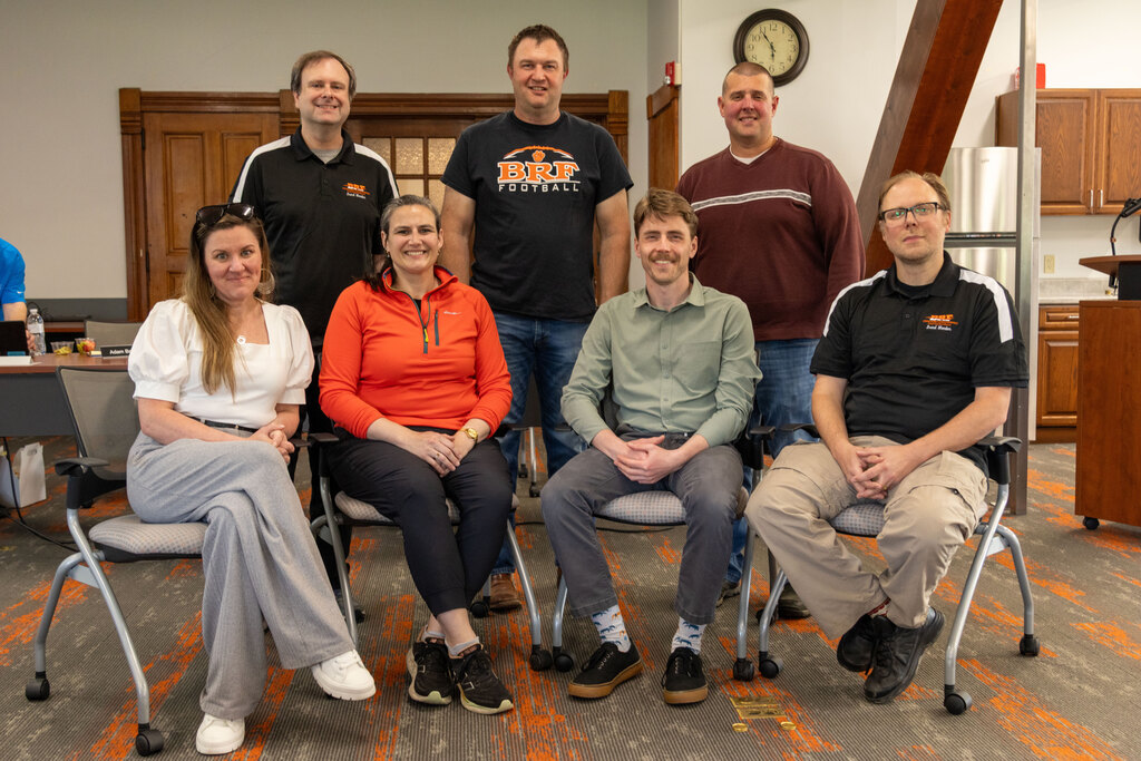 Seven adults, four seated in front and three standing behind them, pose together in a room for a group photo.