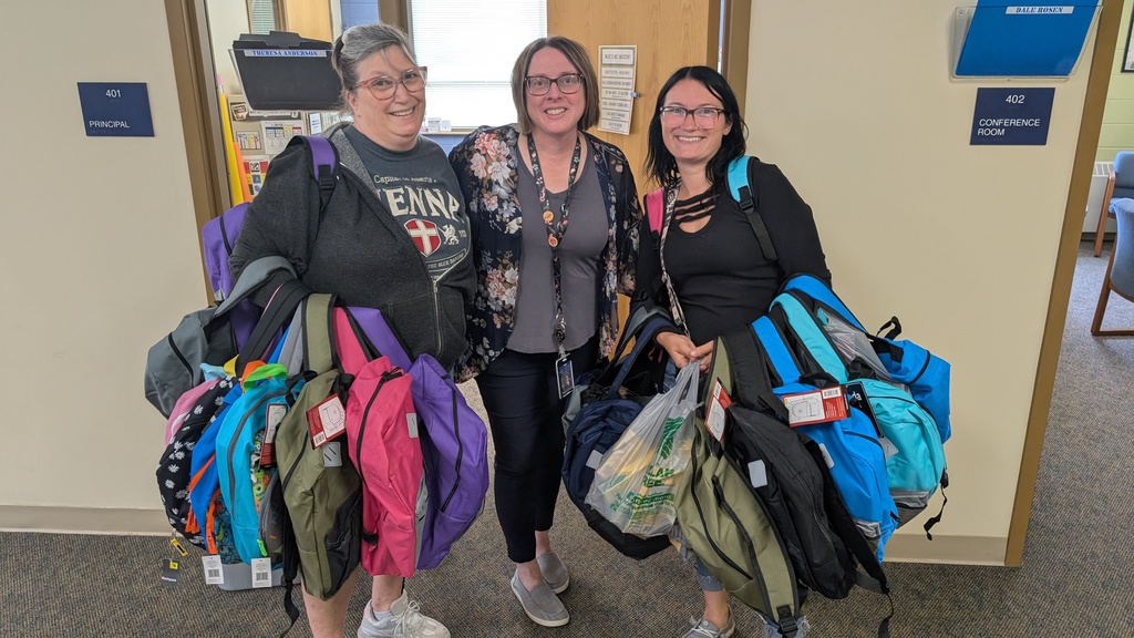3 ladies smiling and holding arms full of backpacks