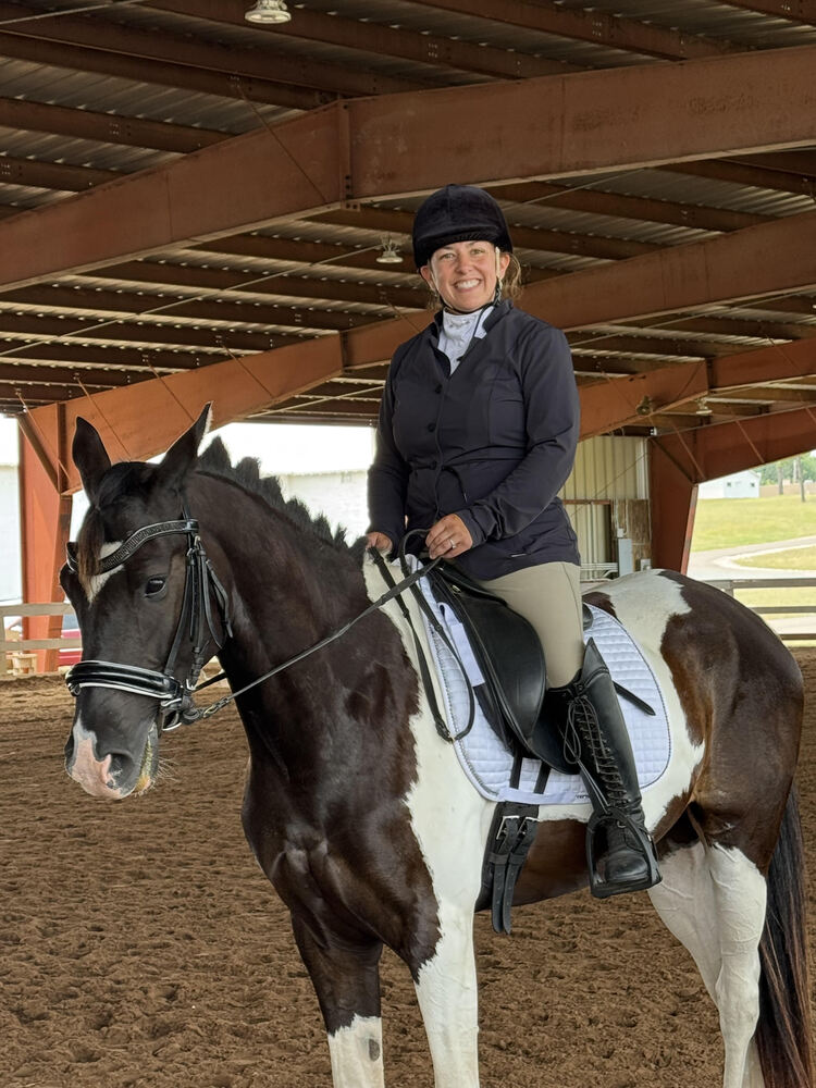 A smiling woman wearing equestrian riding gear and a helmet, riding a black and white pinto horse inside a covered arena.