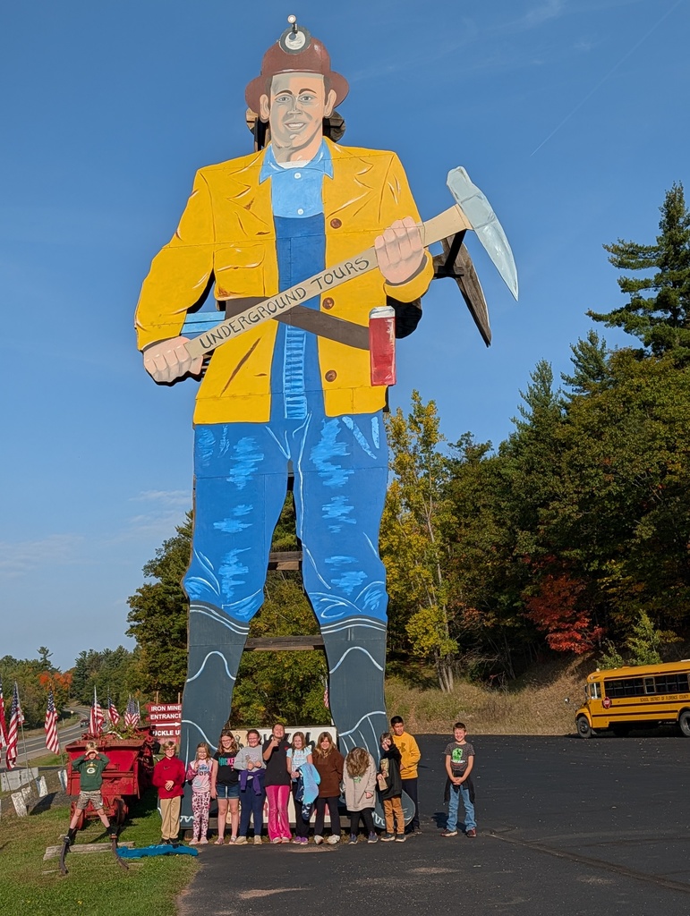 A group of 4th grade students stand smiling at the base of a towering miner statue at the Iron Mountain Iron Mine. The miner wears a yellow jacket, blue overalls, and a red hard hat with a headlamp. He holds a pickaxe and a sign reading “Underground Tours.” A school bus is parked to the right, and American flags line the roadside on the left. The trees in the background show early fall colors.