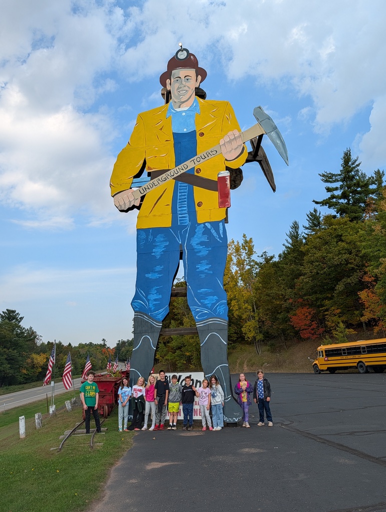 Another group of 4th grade students poses beneath the same giant miner statue at the Iron Mountain Iron Mine. The children stand in a row across the statue’s legs, with a bright blue sky above and autumn trees in the background. A yellow school bus is parked on the right side, and American flags are displayed along the roadside on the left.