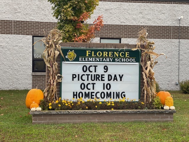 A decorative outdoor sign in front of Florence Elementary School announces upcoming events. The sign reads: “OCT 9 PICTURE DAY” and “OCT 10 HOMECOMING.” It is framed with cornstalks, pumpkins, and yellow flowers, set against a brick and white building with a window in the background and autumn-colored leaves visible above the sign.