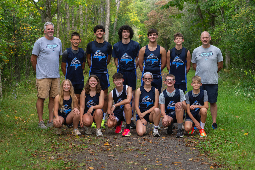 A group photo of the Florence/Niagara cross country team taken outdoors on a wooded trail. Two adult coaches stand on each end, while the team members, wearing navy blue and light blue “Storm” uniforms, are arranged in two rows—six athletes standing in the back and five kneeling in front. The team is smiling and surrounded by lush green trees and early autumn foliage, creating a bright and natural backdrop
