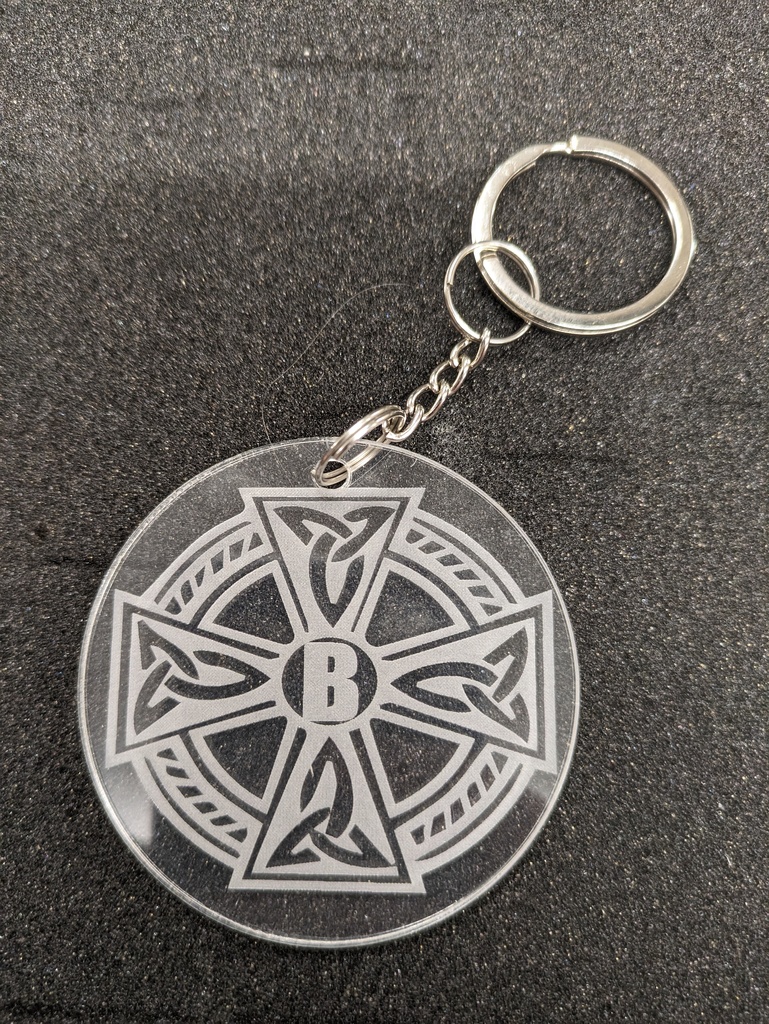 Alt text: “A round clear acrylic keychain with a silver key ring and chain, featuring a laser-etched Celtic-style cross design with interwoven patterns. The letter ‘B’ is engraved in the center of the cross, all set against a black textured background.”