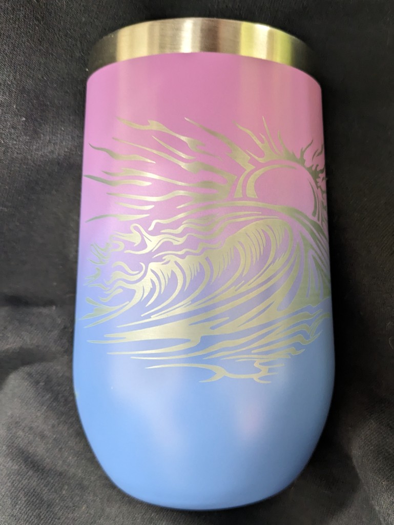 Alt text: “A stainless steel tumbler with a gradient design that fades from pink at the top to blue at the bottom. It features a laser-etched image of a stylized sun and ocean waves, creating a flowing, artistic pattern. The cup rests on a dark fabric background.”