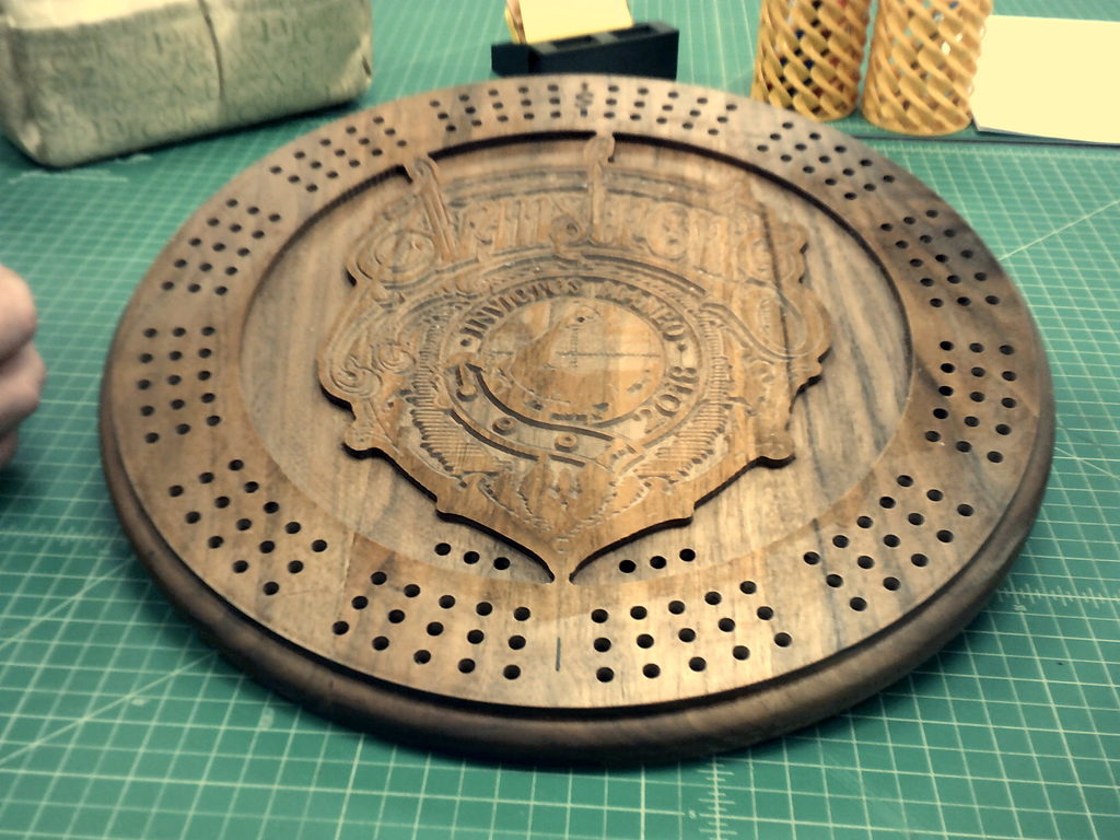 A handcrafted wooden cribbage board featuring an intricate engraved design in the center with decorative lettering and a circular pattern of peg holes around the edge. The board rests on a green cutting mat, with additional wooden pieces and crafting materials visible in the background.”