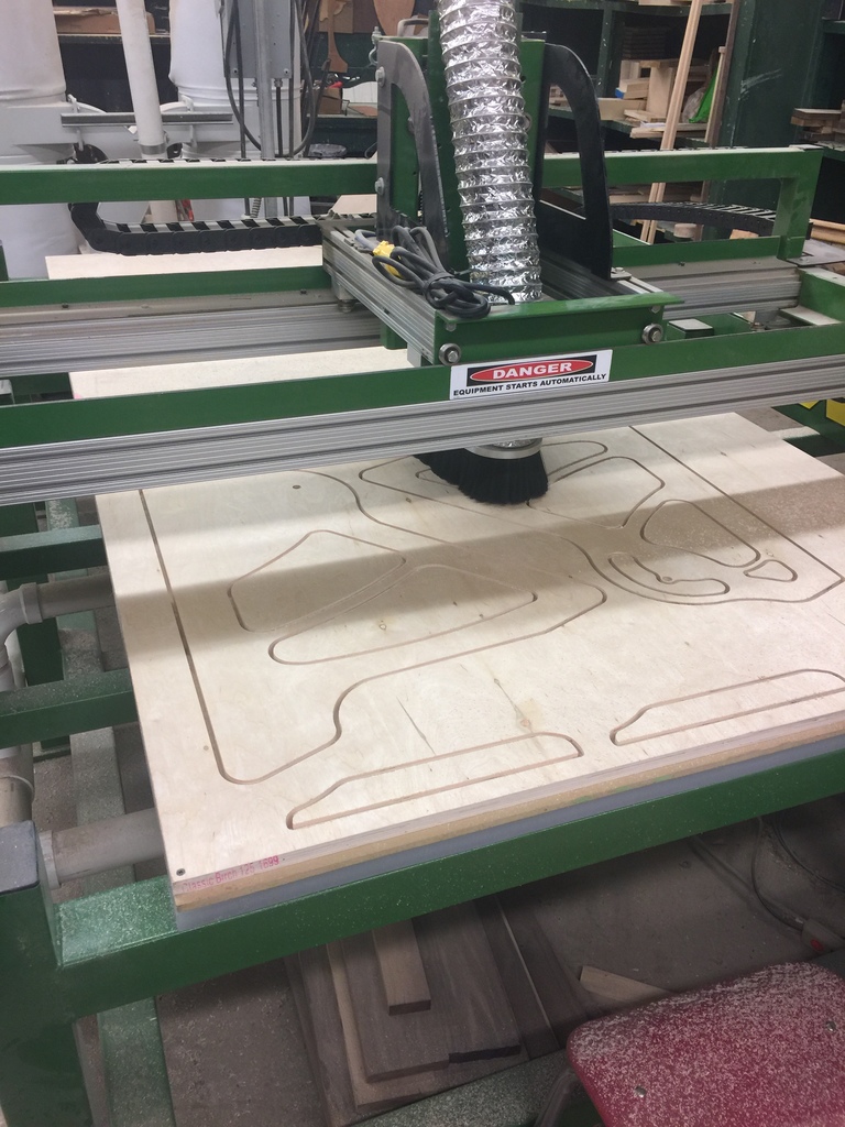 “A CNC router machine cutting shapes into a sheet of plywood in a workshop. The green metal machine has a dust collection hose attached and a red warning label that reads ‘Danger: Equipment starts automatically.’ Wood dust and tools are visible around the workspace.”