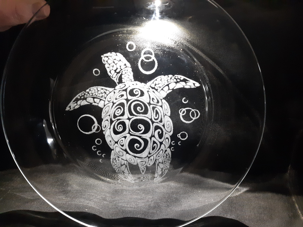 Alt text: “A clear glass plate featuring a detailed etched design of a sea turtle with a spiral pattern on its shell, surrounded by etched bubbles. The plate is placed against a dark background, highlighting the intricate laser-engraved artwork.”
