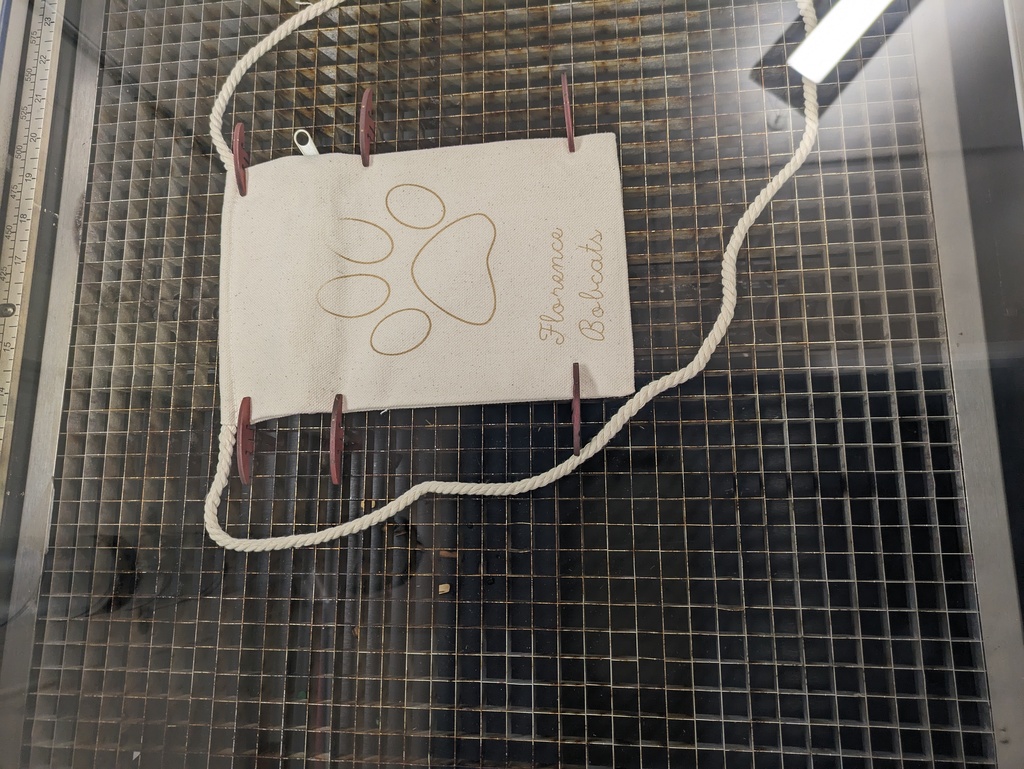 Alt text: “A small canvas bag with a rope handle secured inside a laser engraving machine. The bag is being etched with a design featuring a large paw print and the words ‘Florence Bobcats’ in script lettering.”