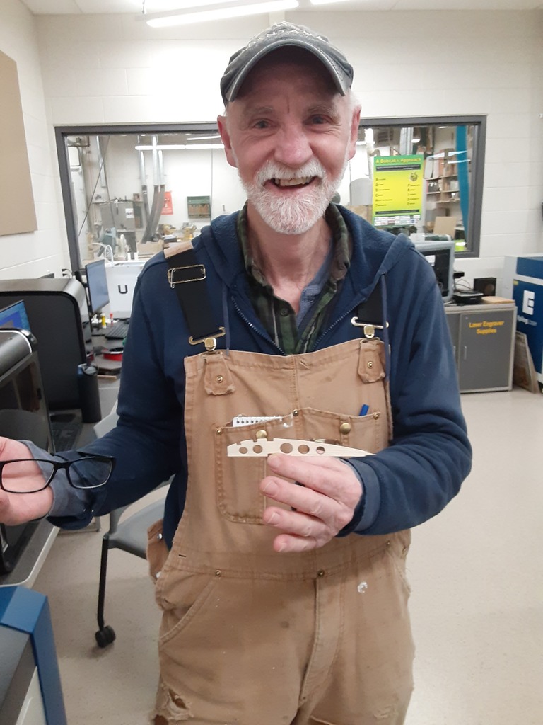 Alt text: “A smiling man wearing tan overalls and a blue cap stands in a workshop, holding a small wooden model airplane wing rib with circular cutouts. The background shows equipment and tools inside the Florence Fab Lab.”