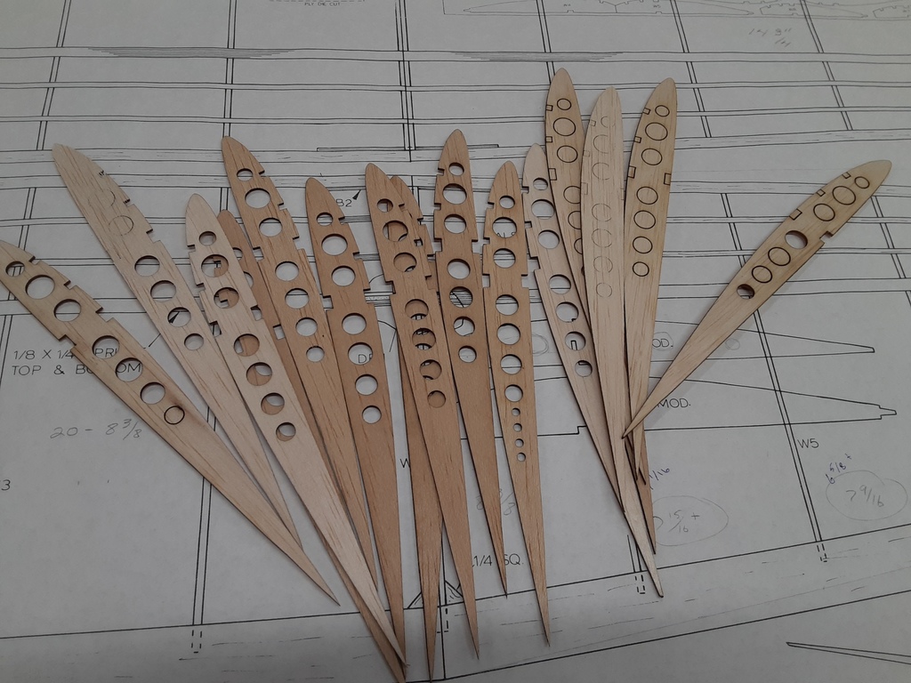 Alt text: “Several thin wooden model airplane ribs with precision-cut circular holes are arranged in a fan shape on top of detailed blueprint plans for an aircraft wing design.”