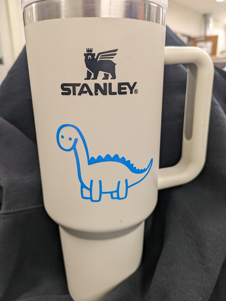 Alt text: “A beige Stanley travel mug with a handle, featuring a cute blue vinyl decal of a smiling cartoon dinosaur with small legs, a long neck, and spiky back. The mug rests on a dark fabric background.”