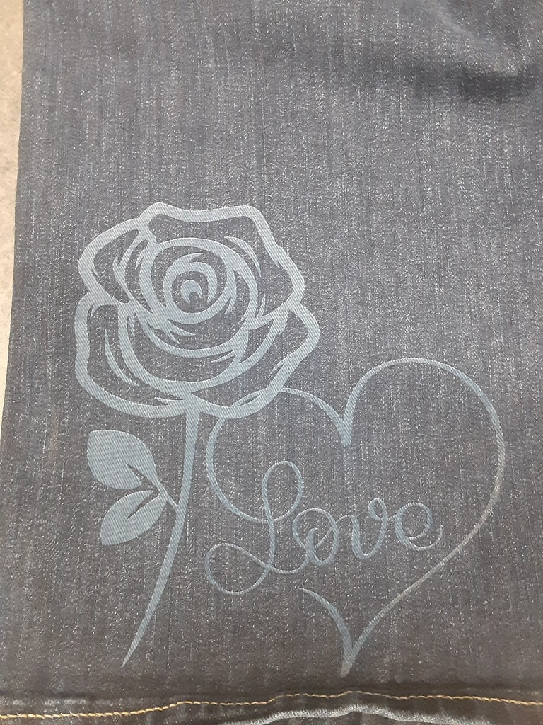 Alt text: “A close-up of denim fabric featuring a laser-etched design of a rose with leaves next to a heart outline containing the word ‘Love’ written in elegant script.”
