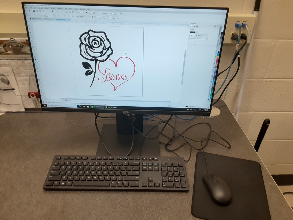 Alt text: “A computer workstation displaying design software with an image of a rose and heart containing the word ‘Love.’ The setup includes a Dell monitor, keyboard, and mouse on a gray desk, with a clipboard and USB drive visible to the left.”