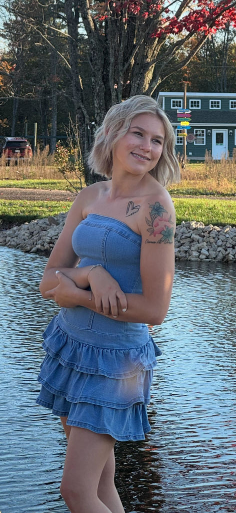 Alt text: “A young woman with short blonde hair stands outdoors near a pond, smiling softly with her arms crossed. She is wearing a light blue denim dress and has colorful tattoos on her shoulder, including flowers, a heart, and cursive writing. Autumn trees and a small green building appear in the background.”