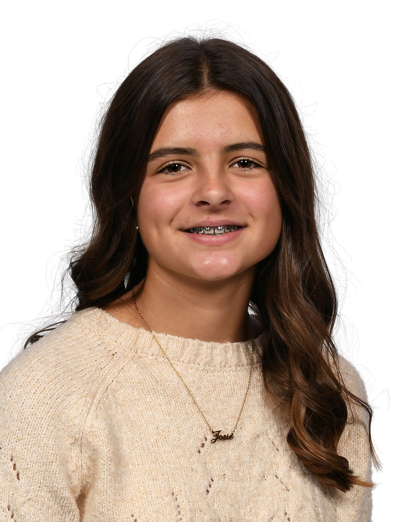Alt text: “A portrait of a young girl with long brown hair and braces, smiling at the camera. She is wearing a cream-colored knit sweater and a gold necklace with a name pendant that says ‘Josie,’ against a plain white background.”