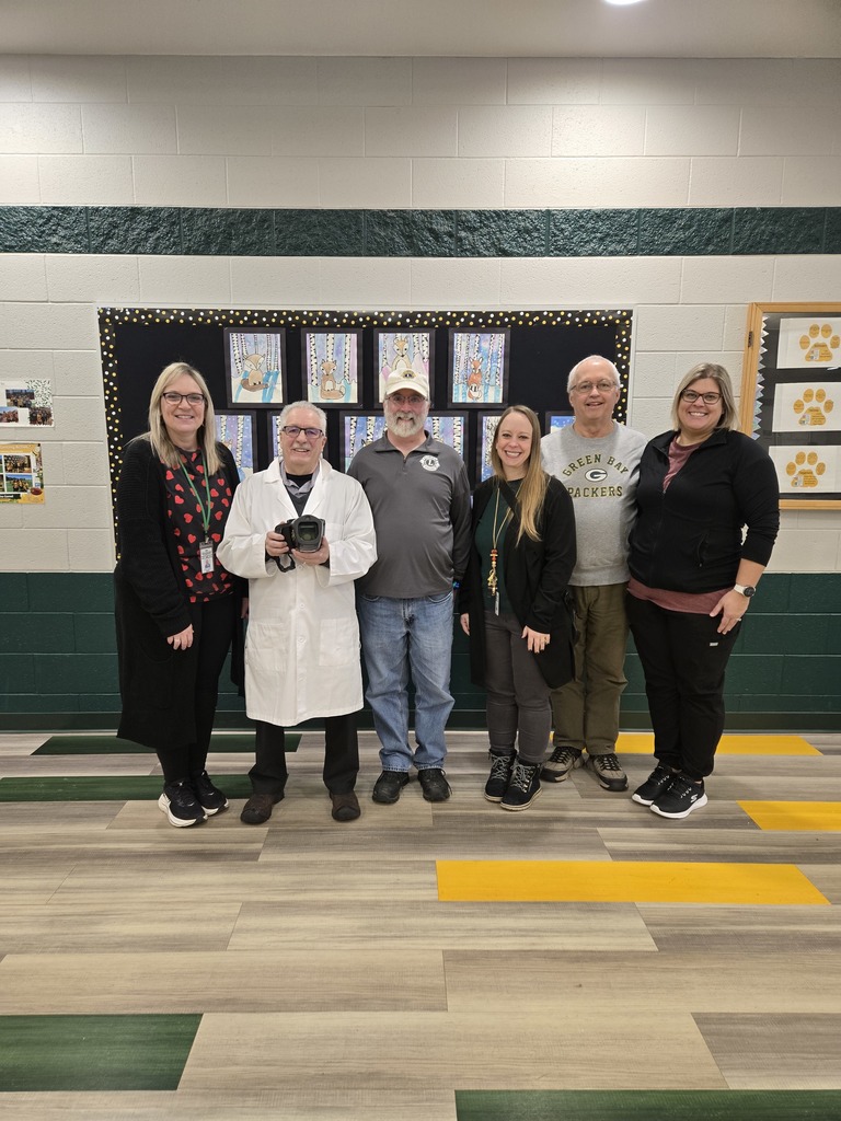 Six adults 3 Florence Elementary School Staff and 3 volunteers from the Lions club stand together in a school hallway, smiling for a group photo. One person in the center is holding a camera and wearing a white lab coat, while the others are dressed in casual or school staff attire, some wearing lanyards. Behind them is a bulletin board displaying student artwork, and the hallway has green and white walls with a patterned floor. The group is the group that administered the Lion's Club Eye screenings to students.
