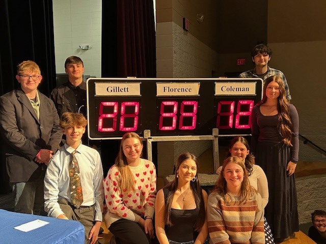 A group of Florence High School students pose around an electronic scoreboard at a Hi-Q academic competition. The display shows scores for Gillett, Florence, and Coleman. Students are smiling and dressed in semi-formal attire inside an auditorium setting.