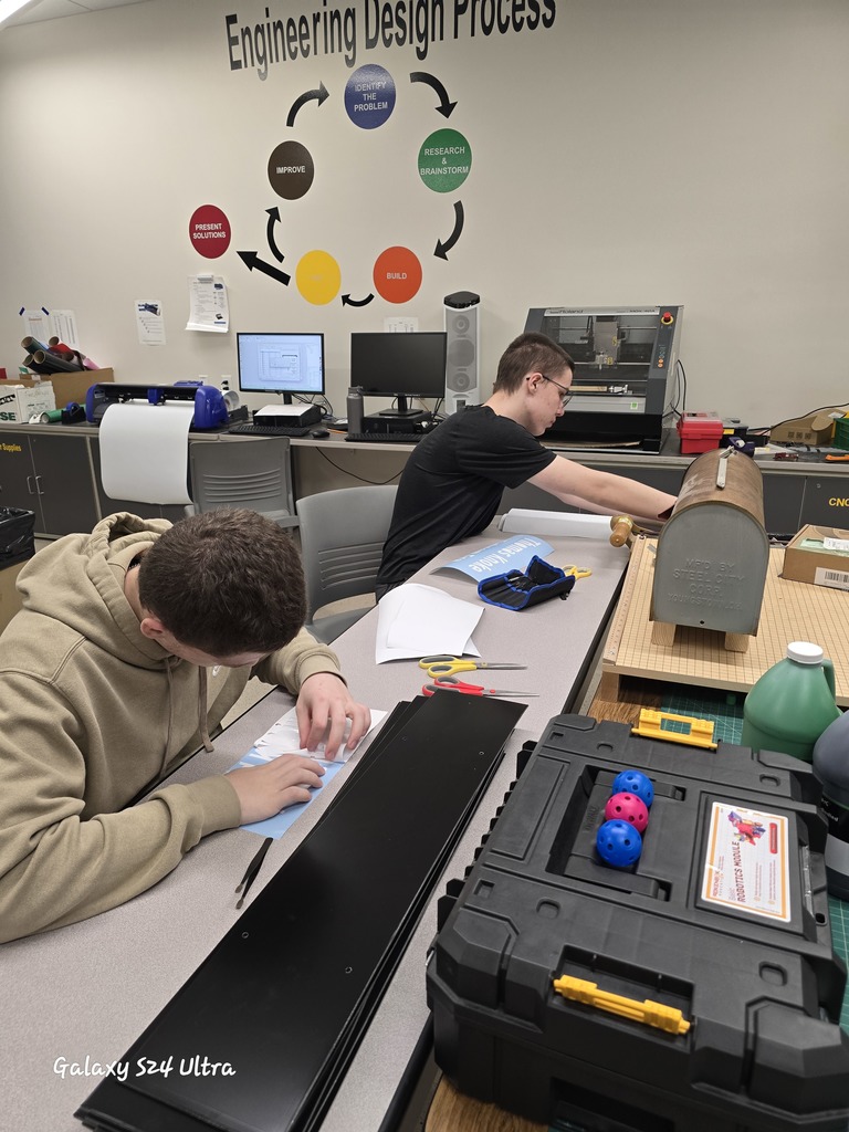 Two students work together in a classroom lab, carefully assembling and applying names to plaques. The Engineering Design Process is displayed on the wall behind them, and tools and materials are spread across the table.