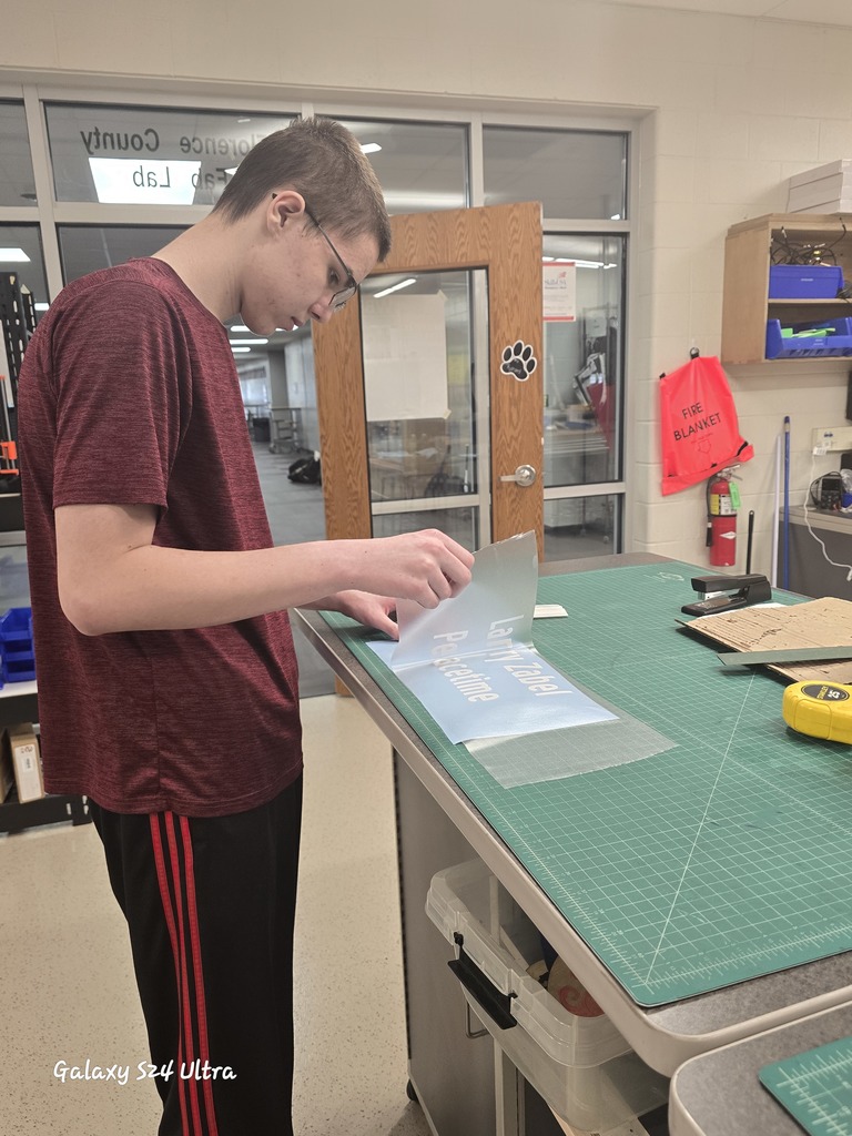 A student carefully applies lettering to a plaque at a worktable, focusing on precise placement. The workspace includes cutting mats, tools, and materials used to prepare Veteran’s Wall name plates.