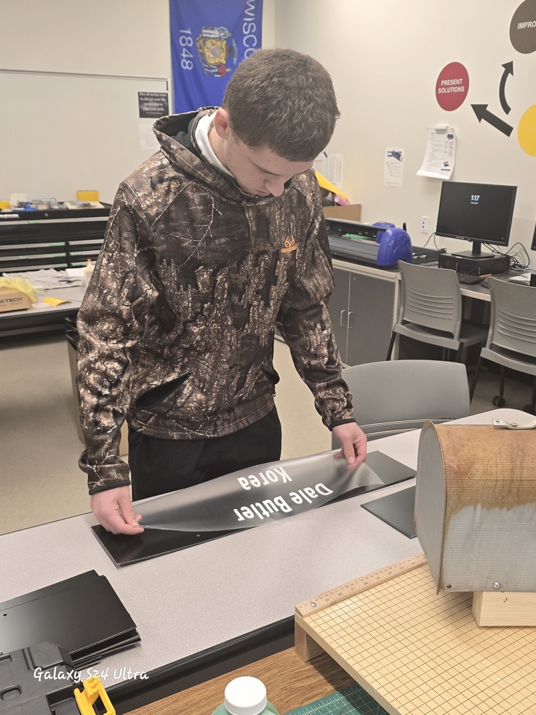 A student aligns and places lettering onto a black plaque, concentrating on spacing and accuracy. A Wisconsin flag and classroom equipment are visible in the background.