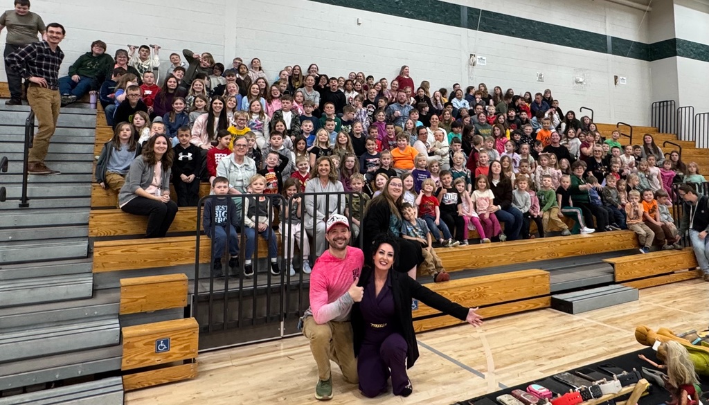 Wide view of Florence Elementary students filling the bleachers in the gym during the presentation, with Ed and Christie at the front and recovered items displayed on a table.