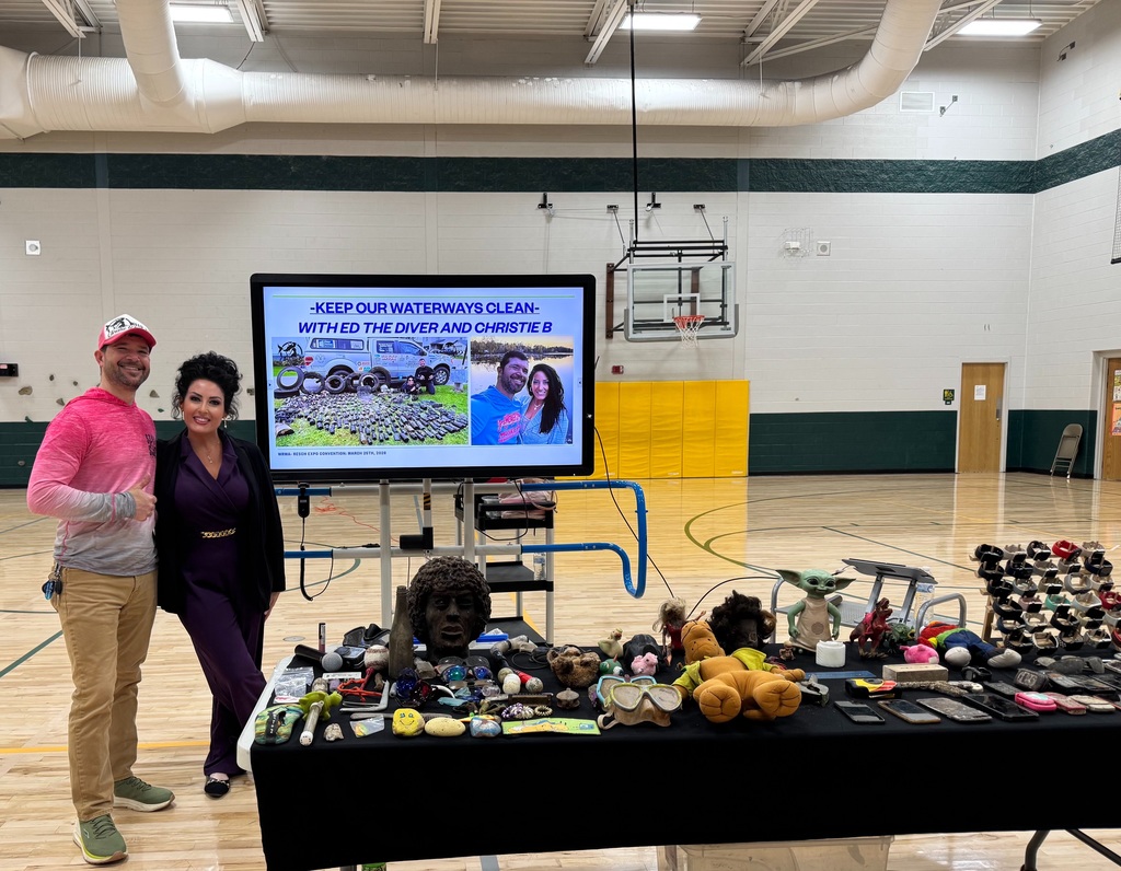 Ed the Diver and Christie B stand in a school gym next to a table filled with items recovered from lakes, including phones, sunglasses, toys, and other objects, with a presentation screen behind them about keeping waterways clean.