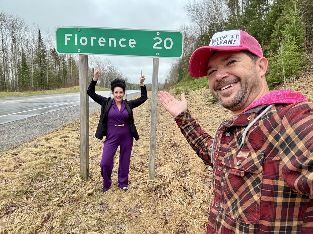 Ed the Diver and Christie B stand by a roadside sign showing the distance to Florence, smiling and pointing at the sign.
