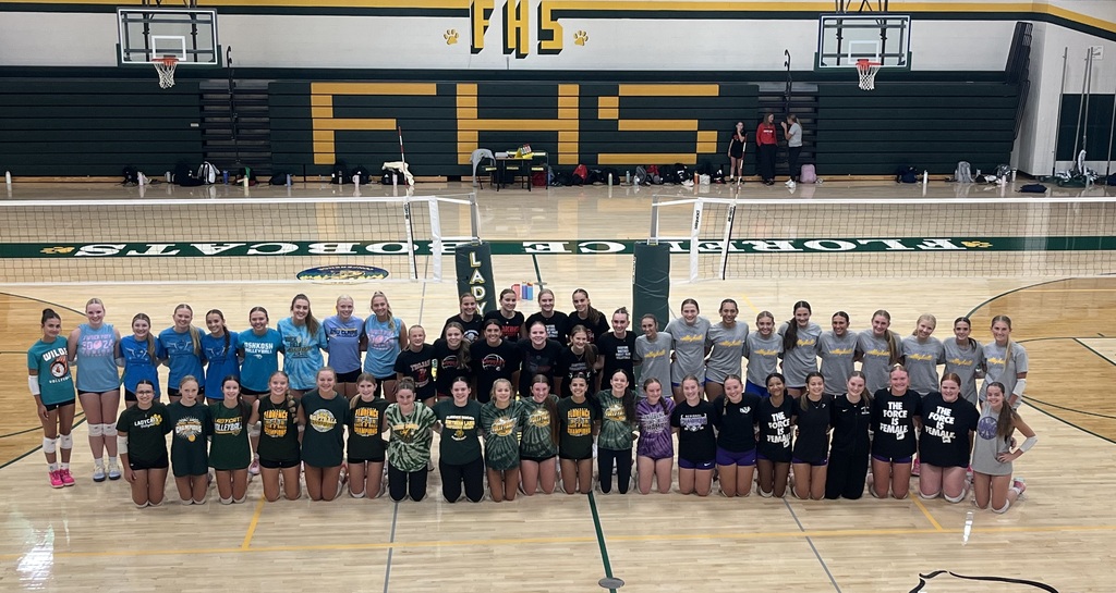 A large group of high school volleyball players from multiple area teams pose together on the court inside the Florence High School gymnasium following a summer volleyball league. The athletes are arranged in three rows in front of the net, wearing a variety of team shirts in green, gray, blue, black, and purple. The gym features green-and-gold Florence Bobcats branding, with large "FHS" lettering on the bleachers. Volleyball nets remain set up behind the group, highlighting the collaborative and competitive atmosphere of the summer league.