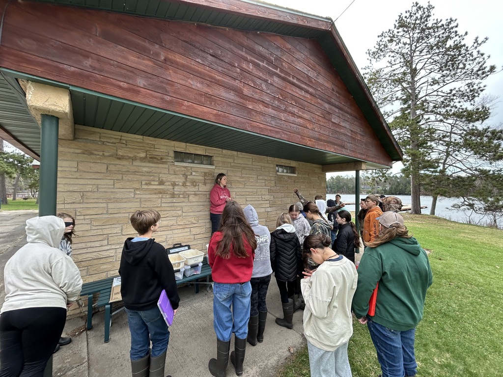 A outdoor educational activity taking place beside a stone-and-wood shelter near a body of water. About a dozen teenagers stand in a semi-circle facing an instructor wearing a red hoodie, who is elevated on a bench against the building's light-colored stone wall. On a nearby bench, plastic storage tubs and supplies are set out. The group is dressed casually in sweatshirts, jackets, jeans, and rain boots. In the background, green grass stretches toward a calm lake bordered by tall pine trees under a cloudy sky.