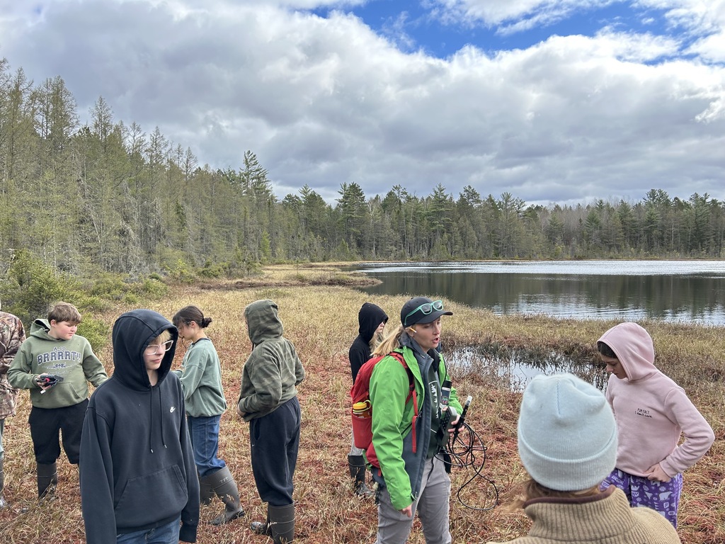 An outdoor field lesson in a wetland ecosystem. A guide wearing a bright green jacket, a backpack, and a baseball cap holds monitoring equipment while addressing a group of children and teens bundled up in hoodies, beanies, and tall waterproof boots. They are standing on the spongy, reddish-brown vegetation of a bog adjacent to a body of calm, dark water. Dense evergreen trees line the distant shore beneath a dynamic sky filled with white clouds and patches of blue.