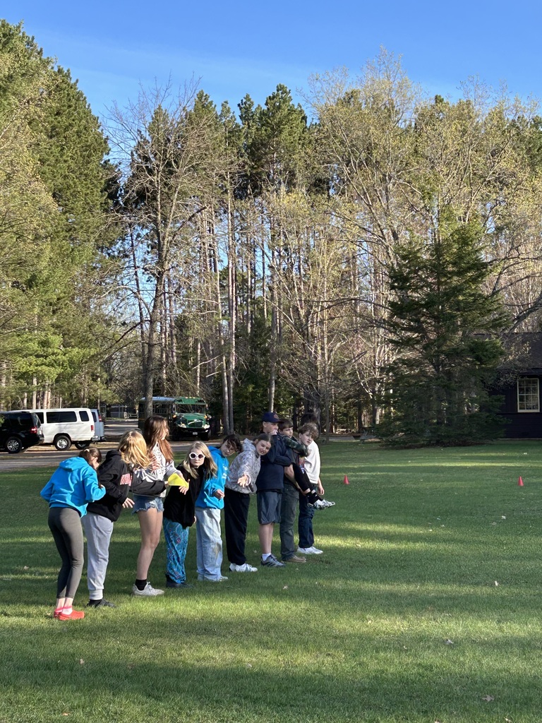 A group of about nine young people participate in an outdoor activity on a grassy field surrounded by dense pine and deciduous trees under a clear blue sky. They stand in a straight line facing right, each leaning out to their left to pose for the camera. Several wear casual sweatshirts, hoodies, jeans, and sneakers. In the background behind the trees, a green vehicle, a dark van, and a small building with a window are visible.