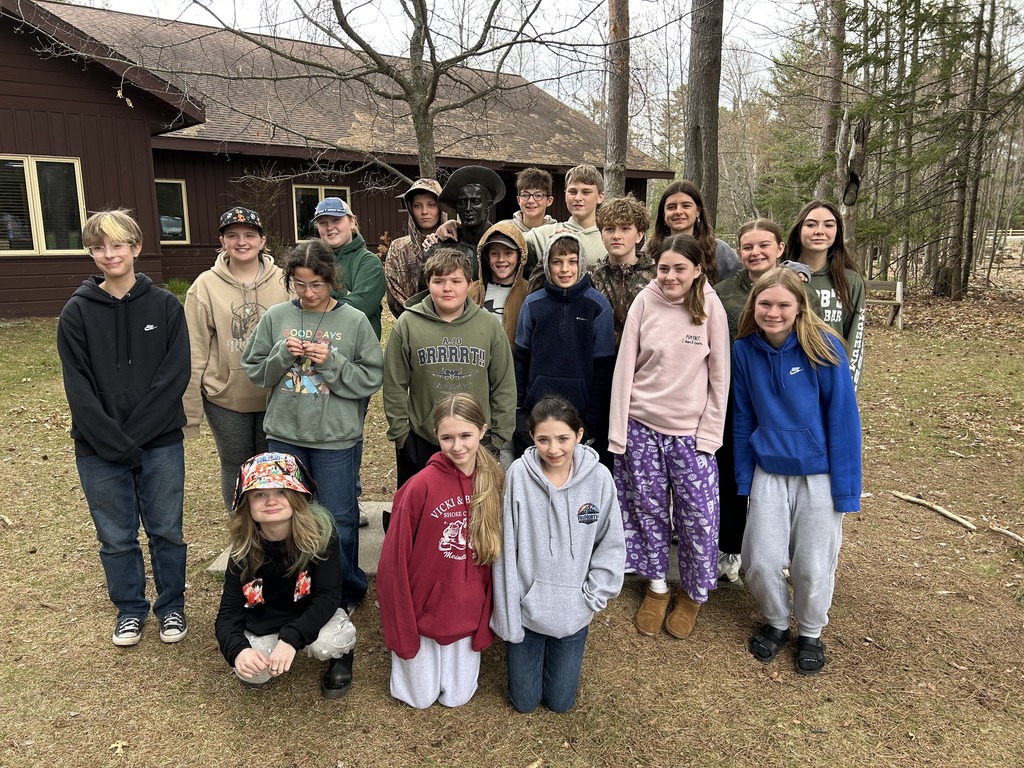 A group photo of students gathered outdoors in a clearing next to a brown wooden building. The children are dressed in various colorful hoodies, sweatshirts, jackets, and hats suitable for cool weather. In the upper middle of the group, a dark metal bust statue of a man with a hat is integrated seamlessly into the gathering, with several kids posed right around it. The group is arranged in three rough rows—some kneeling in front, some standing in the middle, and others in the back—all posing on grass and fallen leaves near a forest border.