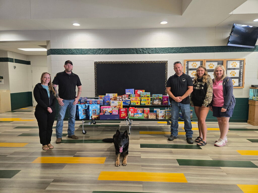 Five people stand in a school hallway around tables filled with donated individually packaged snacks and food items. A black-and-tan German Shepherd lies in front of the tables with its tongue out. The donations include boxes of crackers, granola bars, fruit snacks, applesauce, cereal, and other snacks, along with reusable grocery bags. The group smiles for the photo in front of a bulletin board, highlighting a community effort to support students with snack donations.