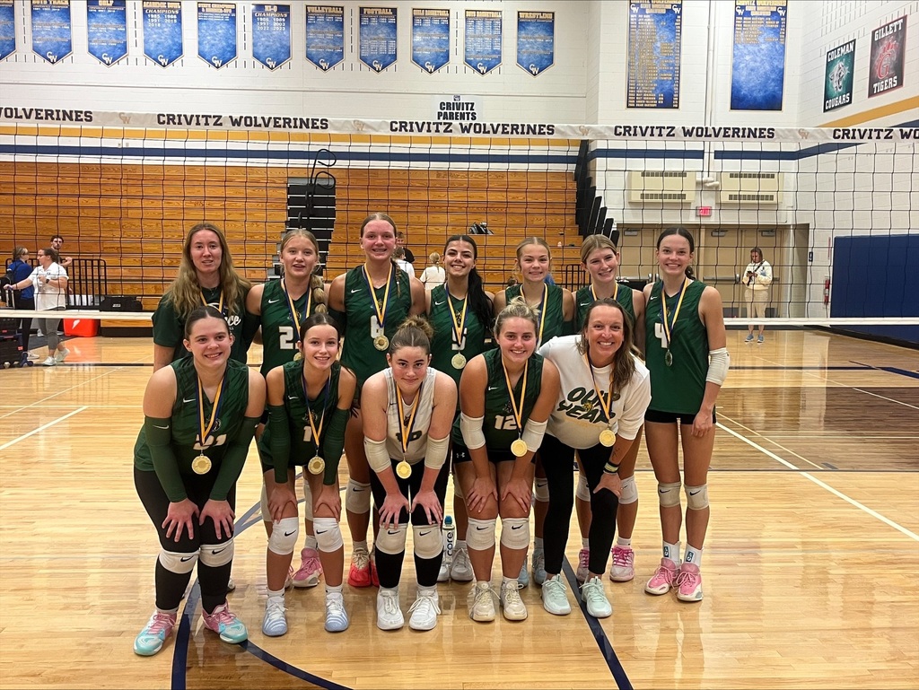 The Florence LadyCats high school volleyball team poses for a group picture inside a gymnasium following a tournament win. Eleven players and two coaches pose in two rows behind a black volleyball net spanning the wooden court. The players wear dark green sleeveless jerseys with gold trim, black shorts, knee pads, and gold medals around their necks. The coach on the far right wears a white sweatshirt that says "OUR YEAR." Banners reading "Crivitz Wolverines" hang on the wall and from the gym structure in the background.