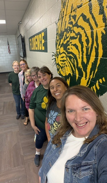 A selfie of a group of seven women lined up diagonally down a brightly lit school hallway with cinderblock walls and wood-look flooring. On the wall beside them is a large painted yellow and dark green mural of a wildcat mascot next to a green sign that reads "FLORENCE" in bold yellow letters. The women are all smiling at the camera, dressed in casual clothing, green tops, and denim jackets.