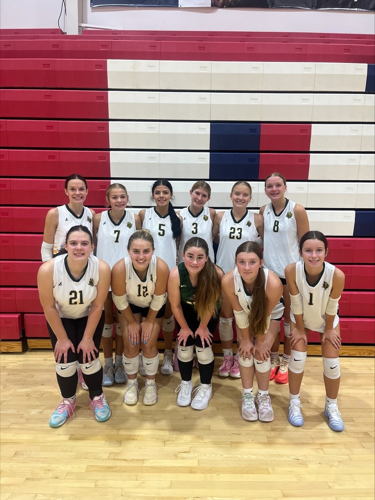 A high school girls' volleyball team poses together on a gymnasium basketball court in front of red, white, and blue bleachers. The eleven players are arranged in two rows—five crouching in the front and six standing behind them. They wear athletic uniforms with white tops, dark shorts, white knee pads, and court sneakers, with the center player in the front row wearing a dark green jersey. Everyone is smiling for the photo.