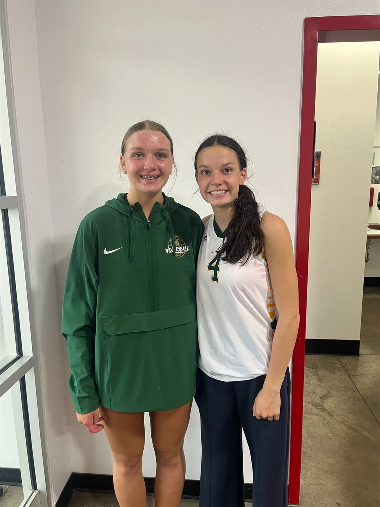 Two high school female volleyball players pose together indoors against a plain off-white wall next to a doorway with a red frame. The player on the left wears a dark green athletic quarter-zip jacket with a volleyball logo and a white Nike swoosh, smiling with braces. The player on the right wears a white volleyball jersey with the number 4 in gold and dark pants, smiling with her arm behind her teammate.