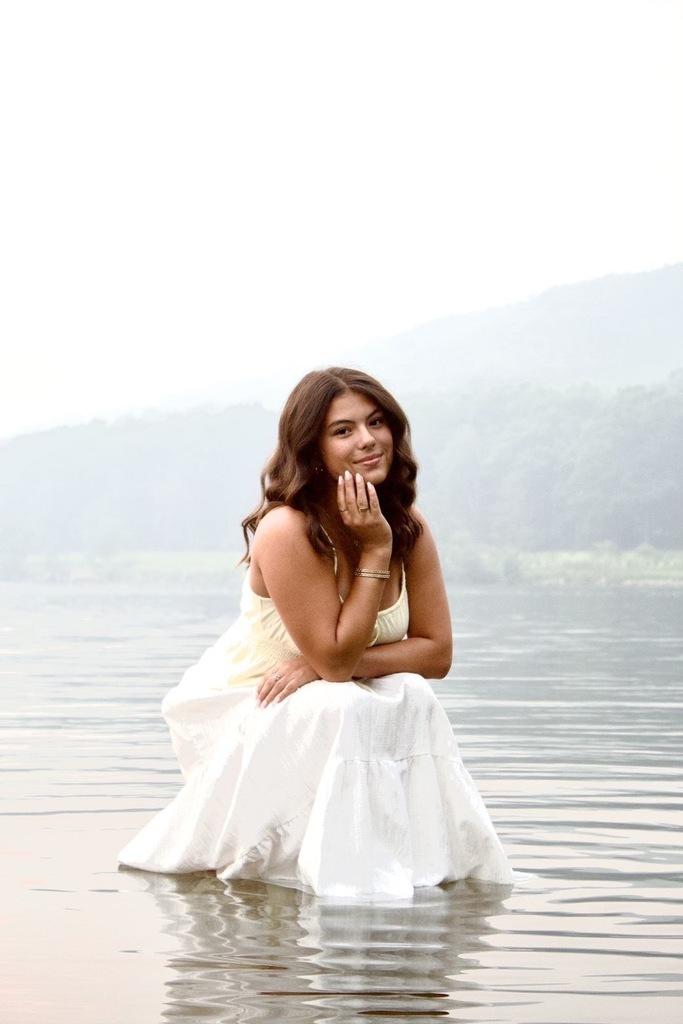 A young woman with shoulder-length wavy brunette hair poses in a tranquil, foggy outdoor lake setting. She is crouching down in the shallow edge of the water, with the lower hem of her long white summer dress resting gently on the surface. She rests one hand lightly against her face near her jawline, looking at the camera with a soft smile. The background features calm, rippling water and hazy, mist-covered hills under a bright, overcast sky.