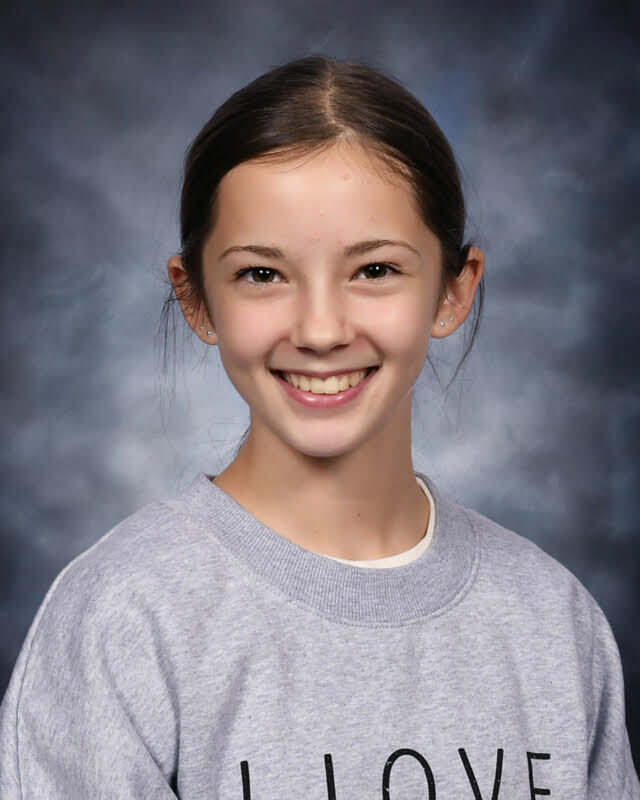 A school portrait of a young girl with dark hair pulled back into a sleek ponytail, smiling brightly in a grey sweatshirt against a mottled blue-grey background.