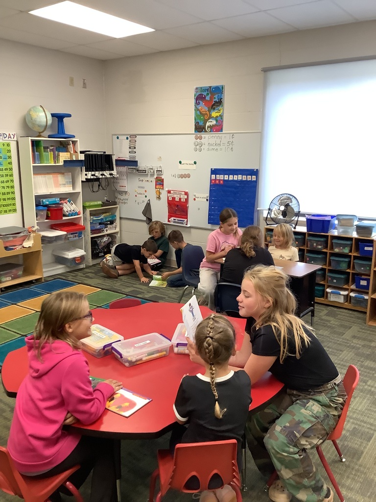A bright, well-organized elementary classroom filled with students working in pairs and small groups. In the foreground, three young girls gather around a round red table while reading and looking at picture books. Other pairs of students are scattered throughout the background—some sitting at small desks and others lying on the carpeted floor reading together. The classroom features bookshelves, whiteboards, and storage cubbies filled with supplies.