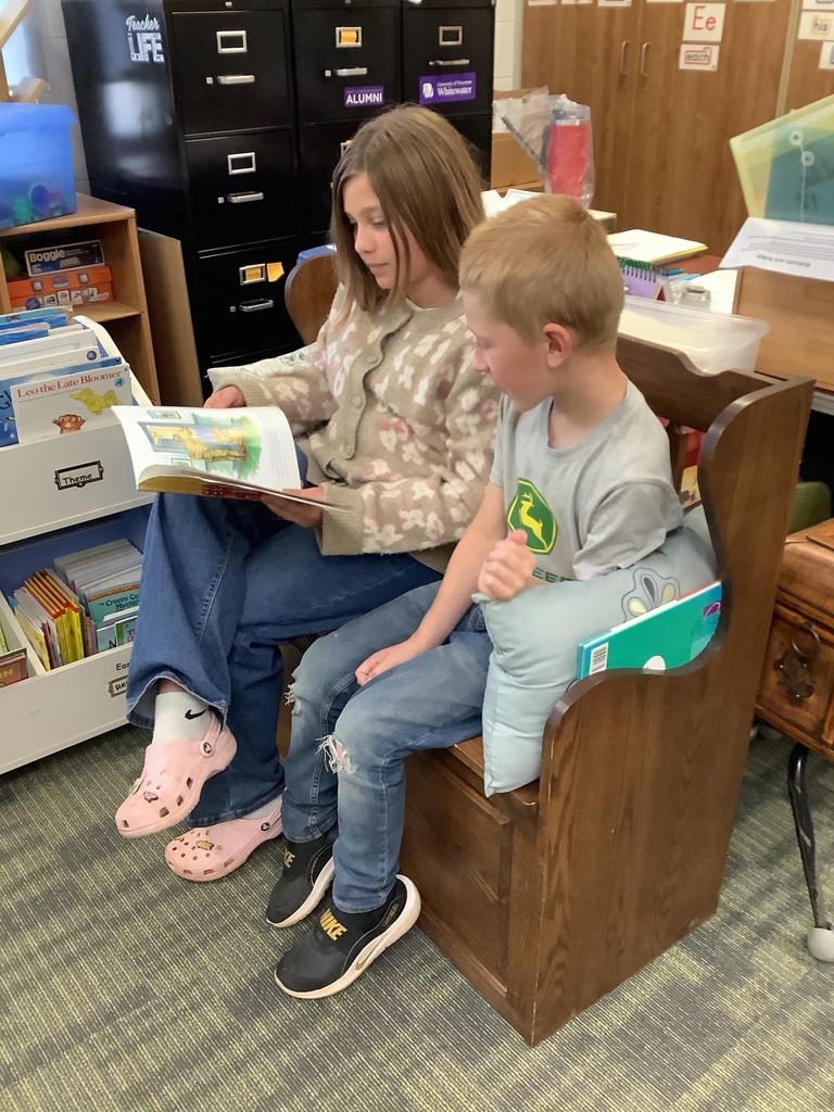 An older student wearing a patterned cardigan and pink Crocs sits next to a younger boy in a grey shirt on a wooden classroom bench, holding open a colorful picture book for them both to read. bookshelf bins and filing cabinets are visible in the background.
